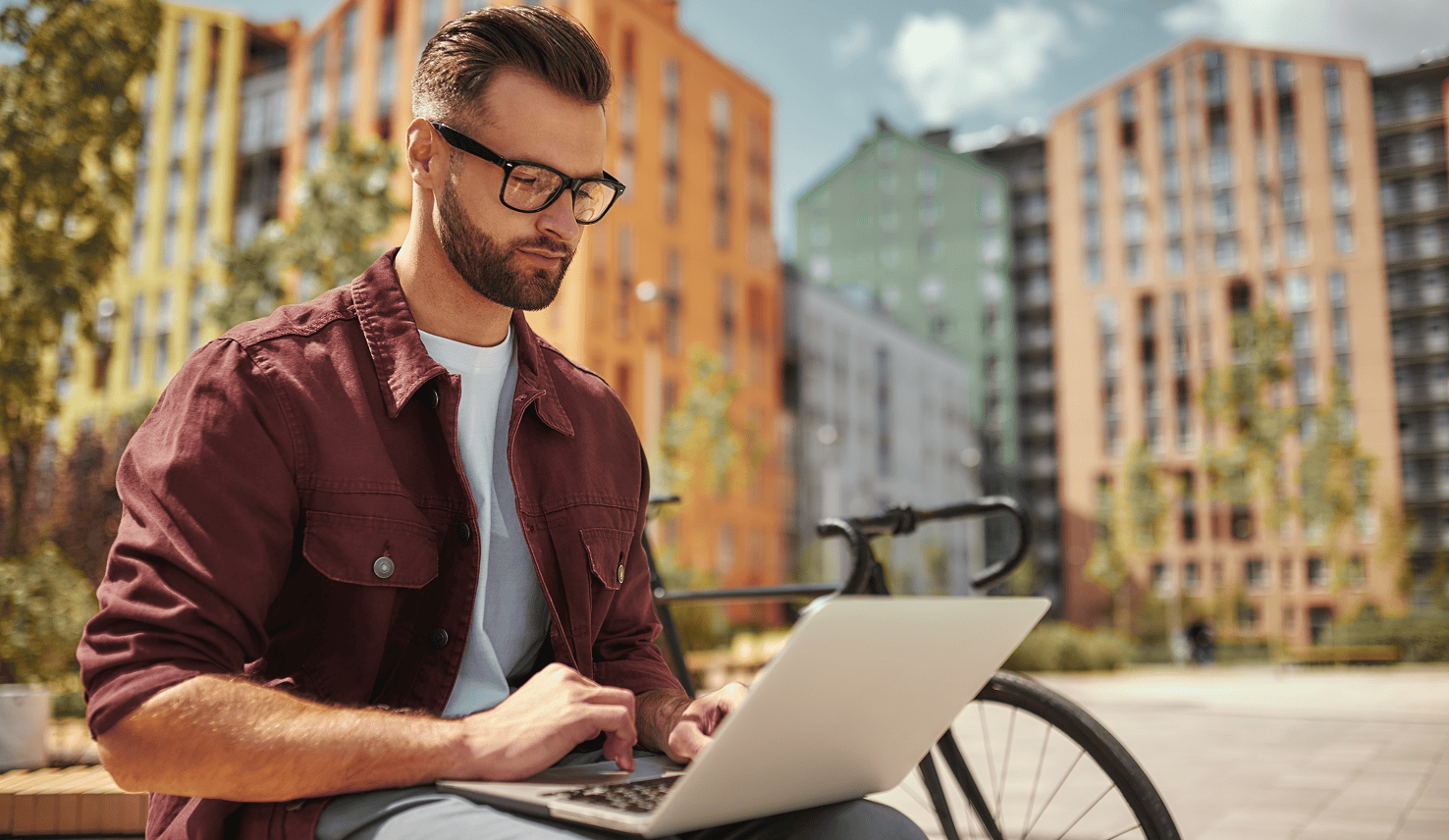 A man working on a laptop outdoors next to his bicycle, with colorful apartment buildings in the background.