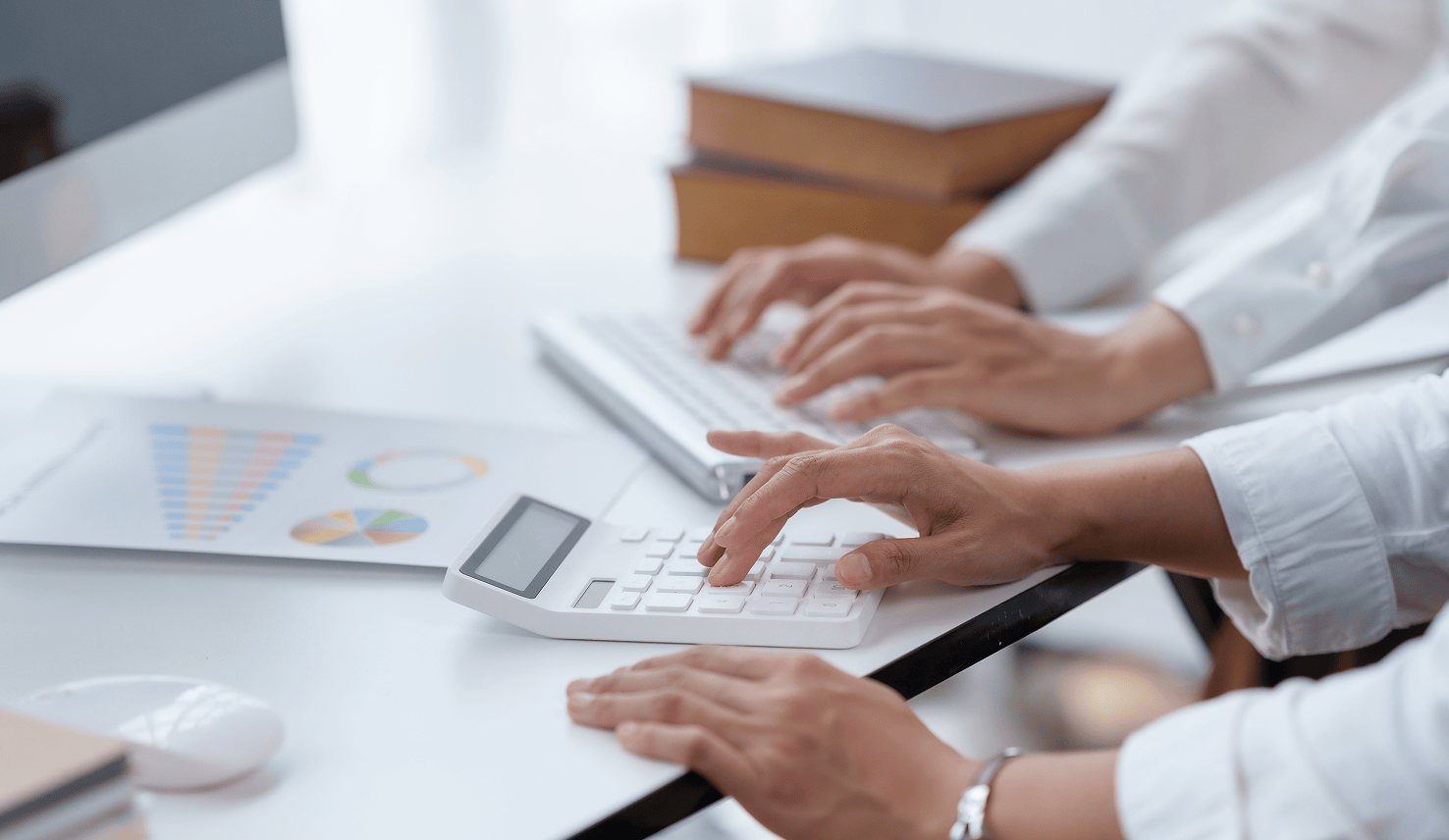 Close-up of two people’s hands wearing white shirts working at a desk in front of a computer, one using a calculator while the other types on a keyboard, with printed charts and graphs nearby.