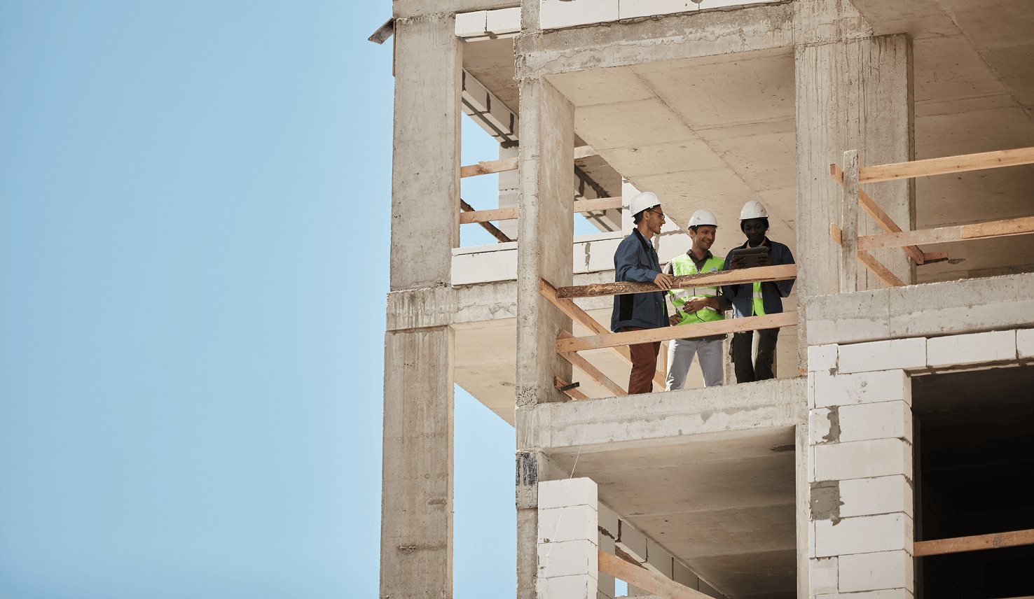 Three construction professionals wearing safety helmets stand on the upper floor of a building under construction, reviewing project plans together.