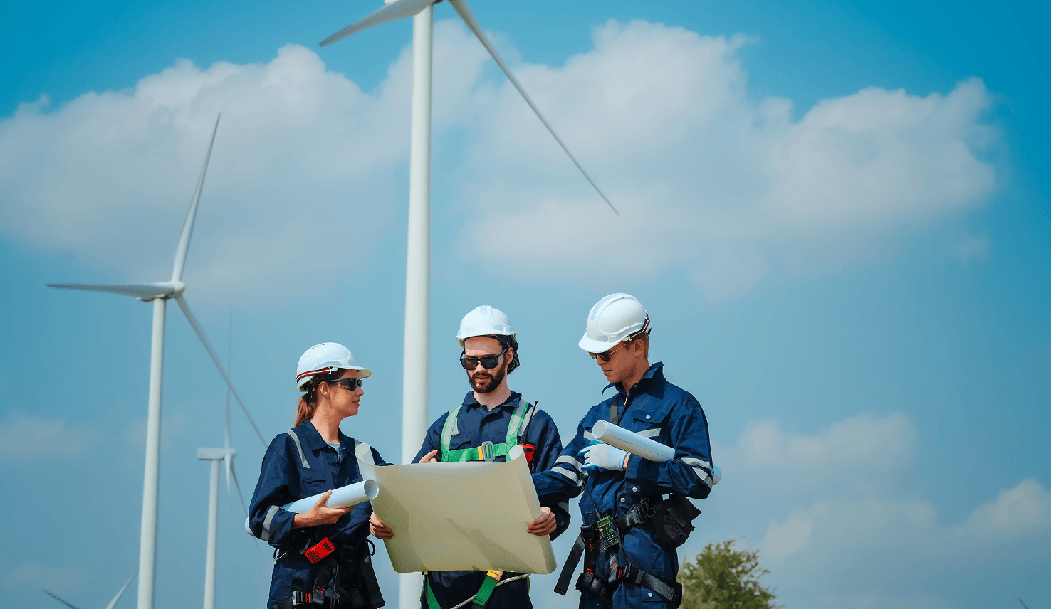 Three wind farm engineers wearing safety helmets review technical plans together in front of wind turbines under a blue sky.