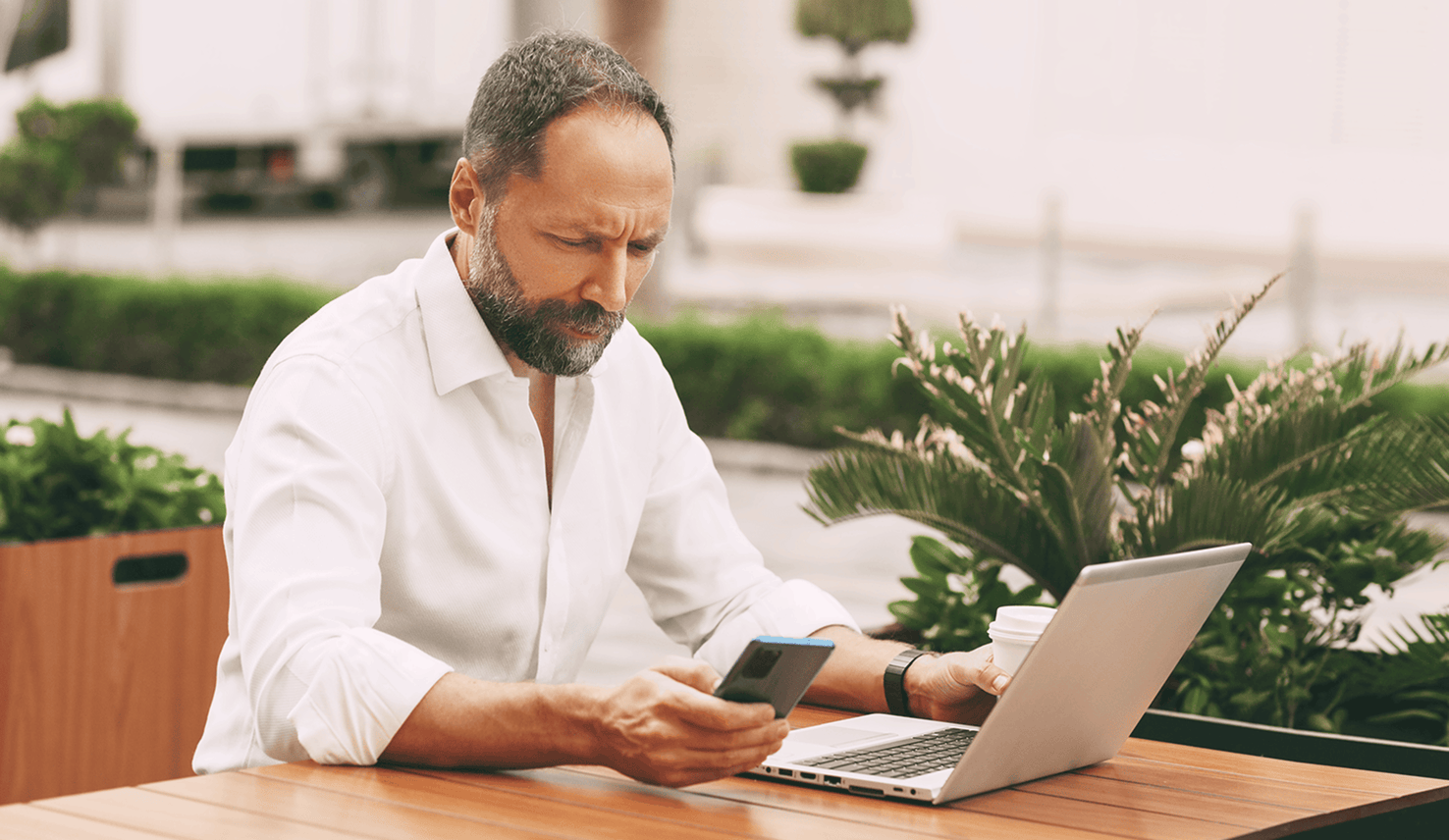Man multitasking on a laptop while checking his phone while sitting at an outdoor café table.
